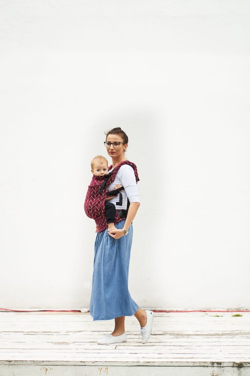 Modelka nosi dziecko w ergonomicznego nosidle KAVKA Multi‑Age burgundy braid cotton - burgundowe nosidło z białym wzorem warkocza  (burgundy carrier with a white braid pattern)
