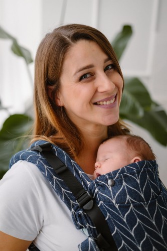 Modelka nosi dziecko w ergonomicznego nosidle KAVKA Multi‑Age ink braid cotton - granatowe nosidło z białym wzorem warkocza (ink braid cotton with white braid pattern)