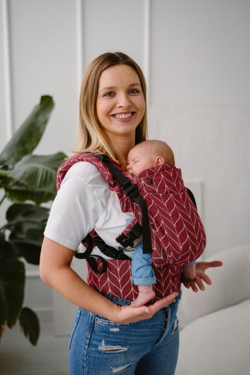Modelka nosi dziecko w ergonomicznego nosidle KAVKA Multi‑Age burgundy braid cotton - burgundowe nosidło z białym wzorem warkocza  (burgundy carrier with a white braid pattern)