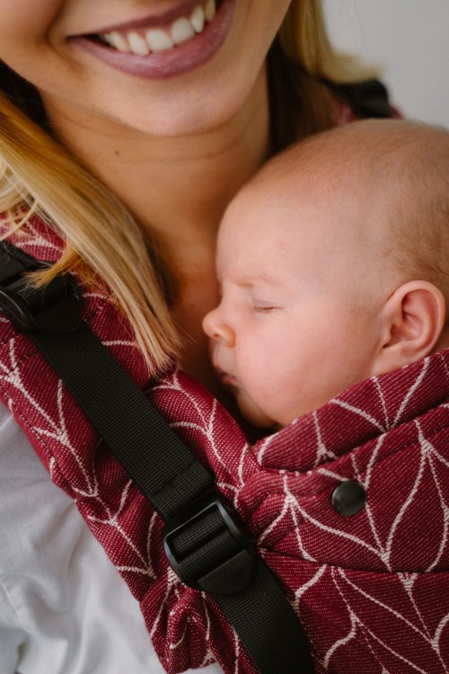 Modelka nosi dziecko w ergonomicznego nosidle KAVKA Multi‑Age burgundy braid cotton - burgundowe nosidło z białym wzorem warkocza (burgundy carrier with a white braid pattern)
