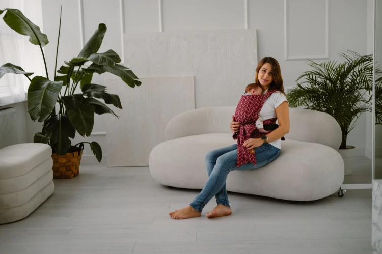 Modelka siedzi na kanapie z dzieckiem w ergonomicznym nosidle KAVKA handy burgundy braid cotton - burgundowe nosidło z białym wzorem warkocza (burgundy carrier with a white braid pattern)