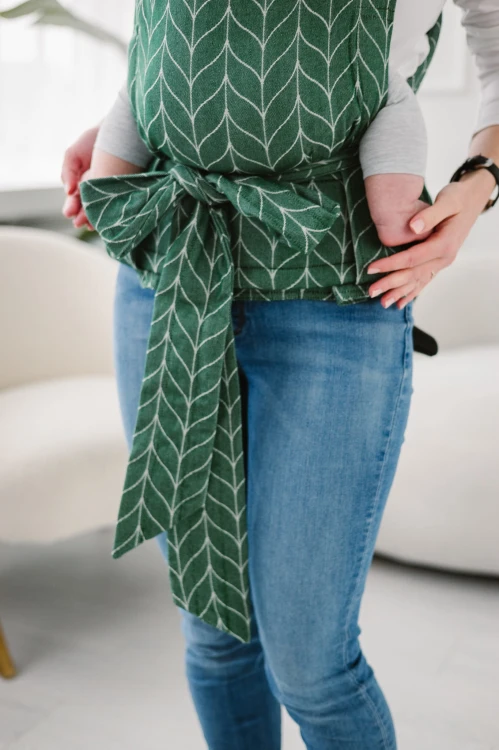 Modelka nosi dziecko w ergonomicznego nosidle KAVKA Handy fern braid bamboo- zielone nosidło z białym wzorem warkocza (green braid  with white braid pattern)